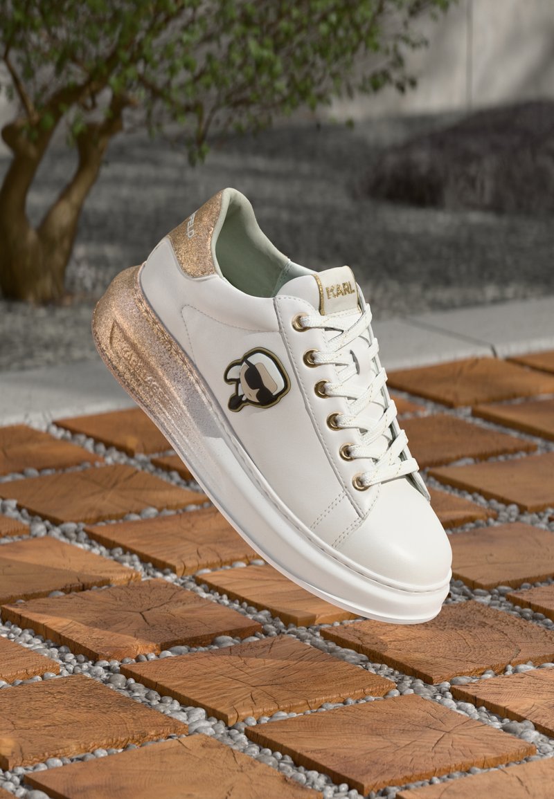 Biały skórzany sneaker z błyszczącym złotym akcentem i logo. Charakteryzuje się przezroczystą podeszwą z teksturowanym wykończeniem. Wyposażony w sznurowanie.