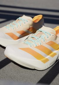 Een paar lichtbeige hardloopschoenen met oranje strepen, lichtblauwe veters en dikke witte zolen op een gestructureerd oppervlak in zonlicht.