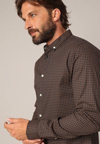 Bruine shirt met patroon en witte knopen, met een klein geometrisch ontwerp en een klassieke kraag. Zachte stof met een soepele textuur.