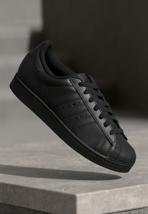 Sort lavprofil sneaker med tekstureret tåhætte, sorte snørebånd og tre sidelængder, vist svævende over en betonoverflade.