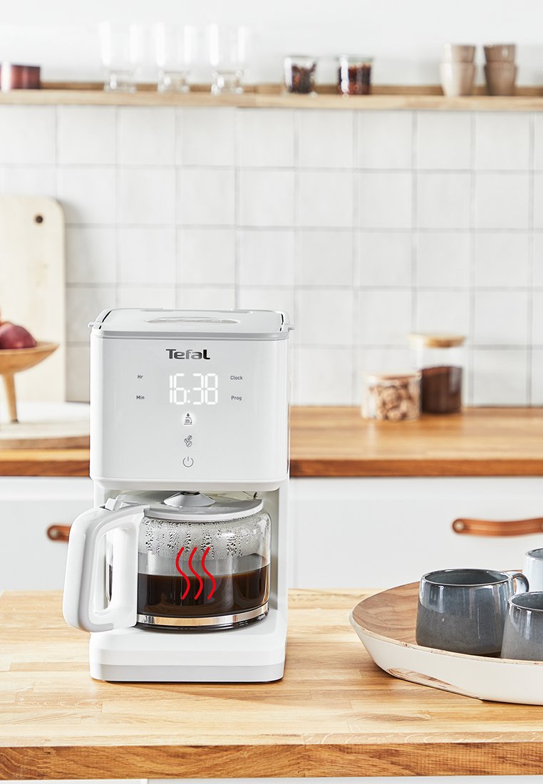 Cafetière blanche avec écran numérique, carafe en verre et indicateur de chaleur visible. Plan de travail en bois avec des tasses grises et un bol en arrière-plan.