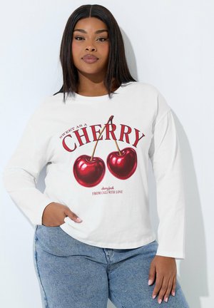 Kvinde med langt mørkt hår iført hvid langærmet trøje med to kirsebær og teksten "Sweet as a Cherry", kombineret med blå jeans.