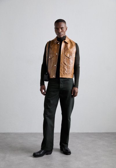 Gilet en cuir synthétique marron avec un col et des poches avant, associé à un pantalon vert foncé et des bottes à talons noires. Sac bandoulière noir simple.