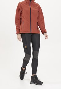 Röd softshell-jacka med hög krage, dragkedjefickor, matchad med svarta leggings med texturerade detaljer och robusta svarta vandringsskor.