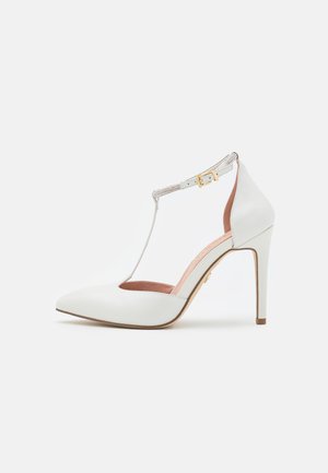 White heeled shoe with a pointed toe, T-strap design, and crystal-accented ankle strap. Smooth leather texture and beige insole.