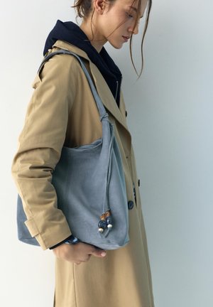 Mladá žena v béžovom trenčkot a čiernej mikine s kapucňou nesie veľkú sivú semišovú kabelku s detailmi z korálikov, stojaca na bielom pozadí.