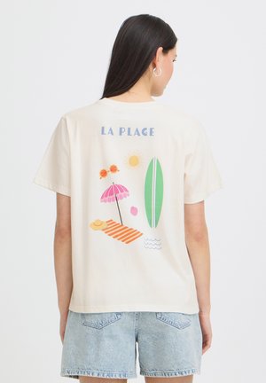 Kobieta o długich ciemnych włosach, ubrana w kremowy t-shirt z nadrukiem w stylu plażowym i napisem „LA PLAGE”, w połączeniu z jasnymi szortami dżinsowymi.