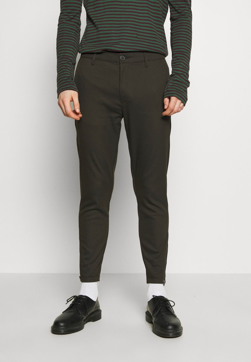 Personne portant un pantalon fuselé vert olive foncé avec des fermetures éclair aux chevilles, des chaussures noires à lacets, des chaussettes blanches et un haut à manches longues rayé vert et noir.
