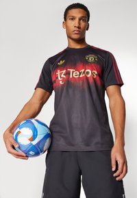 Camiseta de fútbol en degradado negro y rojo con logotipos dorados, detalles rojos y cuello redondo. El modelo sostiene un balón de fútbol con un patrón azul y naranja.