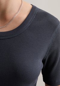 Donkergrijze geribbelde top met korte mouwen en een ronde halslijn, voorzien van zichtbaar stikwerk. Zilveren ketting om de nek.