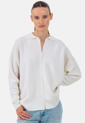 Elfenbeinfarbene, übergroße Strickbluse mit Kragen, durchgehender Knopfleiste und fallenden Schultern, kombiniert mit hellblauen Jeans.