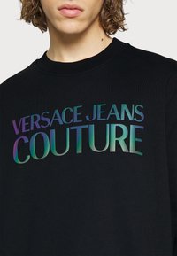 Svart sweatshirt med ribbad krage som pryds av texten "VERSACE JEANS COUTURE" i iriserande gradientfärger. Slät textur, avslappnad design.