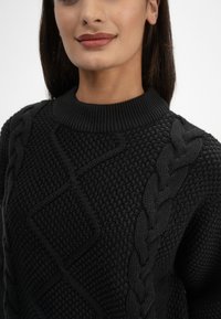 Schwarzer gestrickter Pullover mit strukturierten Mustern, der ein dickes Zopfdesign und einen gerippten Kragen aufweist. Der Stoff wirkt warm und haptisch.