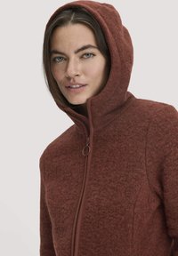Braune Fleecejacke mit einem Reißverschluss vorne und einer Kapuze, ausgestattet mit einem strukturierten Stoff und dezenten Details rund um den Reißverschluss.