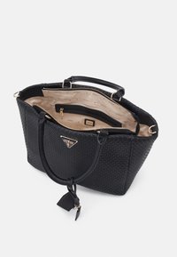 Sac fourre-tout tissé noir avec doubles poignées, doublure intérieure beige, compartiments zippés, et logo triangulaire Guess en métal doré à l'avant.