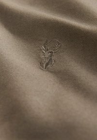 Beige stof met een subtiel reliëf van een hertlogo. De textuur lijkt glad met een strakke weving, wat de duurzame kwaliteit benadrukt.