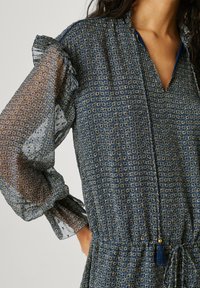 Vrouw die een blauwe jurk met patroon draagt, met doorzichtige blousen mouwen, ruches bij de schouders en een kwastje als strik bij de halslijn.