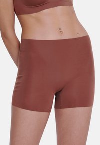 Vrouw in naadloze roestkleurige hoge taille boyshort onderbroek en bijpassende crop top, met een zichtbare navelpiercing.