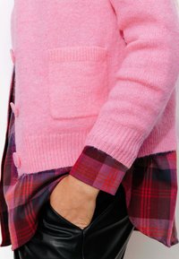 Cardigan en laine rose avec des poignets côtelés visibles et une poche avant, porté par-dessus une chemise en flanelle à carreaux présentant des tons rouges et violets.