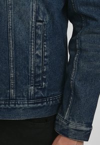 Tumši zila džinsa jaka ar strukturētu dizainu, izceltām šuvēm, krūšu kabatu un salocītām aprocēm. Teksturēta auduma apdare.