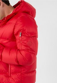 Giacca a vento rossa con cappuccio, dotata di tasche con cerniera, logo in silicone sul braccio e una texture liscia e lucida.