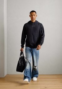 Sort hætte sweatshirt, slidte blå jeans, hvide sneakers og en sort taske. Modellen står foran en lys væg på et trægulv.