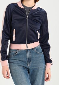 Kvinna i marinblå bomberjacka med rosa detaljer och blå jeans, som står emot en enfärgad ljus bakgrund.