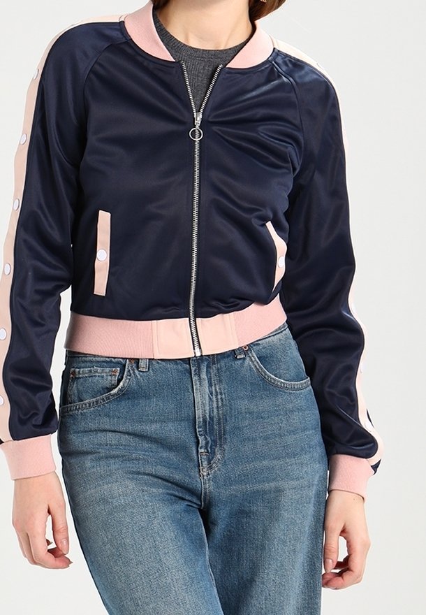 Kvinna i marinblå bomberjacka med rosa detaljer och blå jeans, som står emot en enfärgad ljus bakgrund.