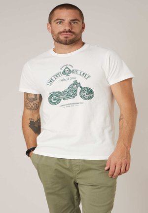 Homme aux cheveux courts et barbe portant un T-shirt blanc avec un motif de moto verte et le texte "Live Fast Die Last", accompagné d'un pantalon olive.