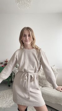 Junge Frau in einem beigen Strickkleid mit Taillenschnürung, stehend in einem Innenraum mit einem Sofa in neutralen Farbtönen, flauschigen Kissen und einem grünen Beistelltisch im Hintergrund.