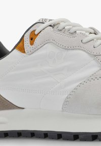 Zapatilla atlética blanca y gris con materiales de ante y sintéticos, que presenta un diseño de calavera y huesos cruzados y acentos en naranja contrastante. Suela de goma.