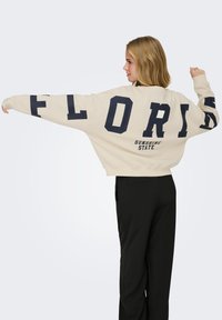 Femme aux cheveux blonds portant un sweat-shirt crème avec les mots "FLORIDE" et "ÉTAT DU SOLEIL" en lettres noires, associée à un pantalon noir, étirant les bras.