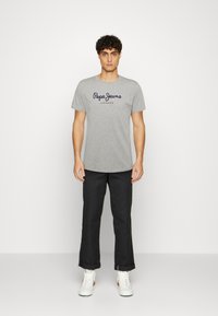 Pilkas medvilninis marškinėliai su "Pepe Jeans London" užrašu tamsiai mėlynos ir raudonos spalvos, dėvimi kartu su juodais kelnais ir baltais sportbačiais, stovintis ant neutralios fono.