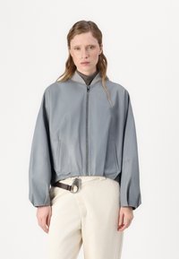 Casaco bomber cinza claro com uma textura suave, ajuste estruturado, gola canelada e bolsos laterais, combinado com calças creme e um cinto.