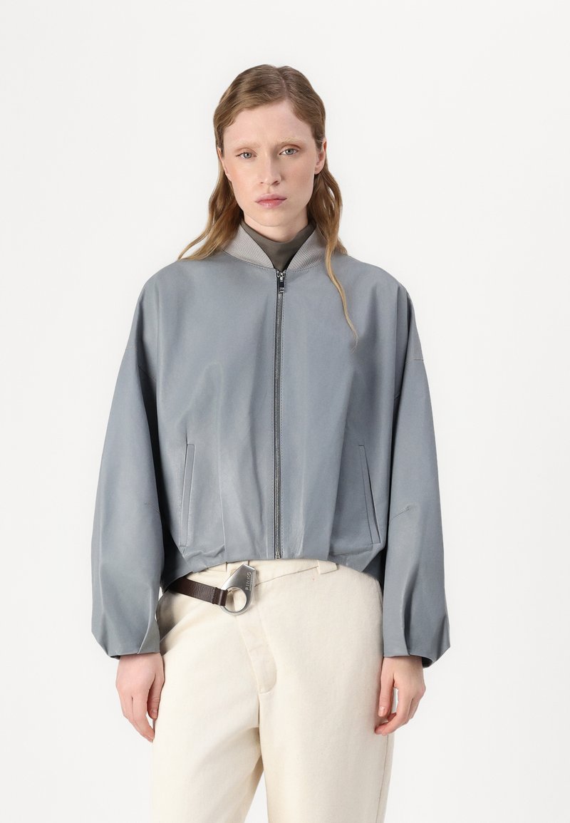 Casaco bomber cinza claro com uma textura suave, ajuste estruturado, gola canelada e bolsos laterais, combinado com calças creme e um cinto.