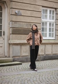 Hnědá texturovaná zipová bunda, šedý šátek, černé kalhoty a tmavé tenisky na pozadí městské krajiny se zdmi a okny.