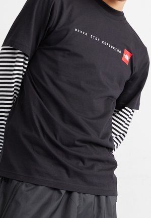Person trägt ein schwarzes T-Shirt mit weißem Text "NEVER STOP EXPLORING" und rotem Quadratslogo über einem gestreiften Langarmshirt und dunkler Hose.