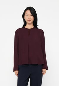 Young woman wearing a long-sleeve burgundy top with a small front keyhole and dark navy pants, standing against a plain white background.
