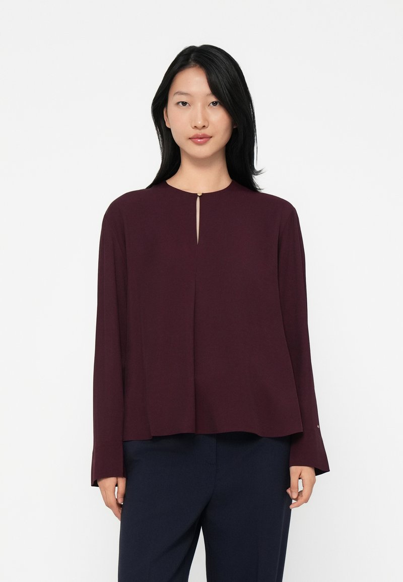 Young woman wearing a long-sleeve burgundy top with a small front keyhole and dark navy pants, standing against a plain white background.