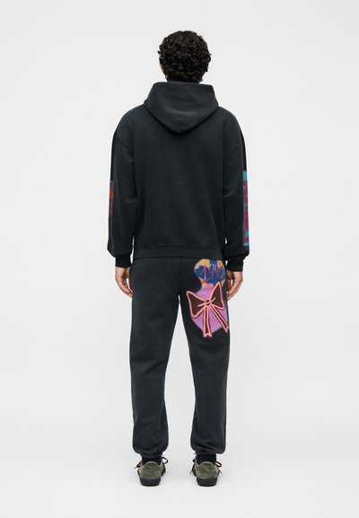 Άνδρας με σγουρά μαλλιά που φοράει μαύρη φούτερ μπλούζα με κουκούλα και φόρμα με πολύχρωμα σχέδια στα μανίκια και στο δεξί πίσω πόδι, στέκεται απέναντι σε λευκό φόντο.