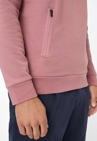 Uomo che indossa un top a maniche lunghe di colore rosa polveroso con una tasca verticale con cerniera e pantaloni blu scuro, con le braccia rilassate lungo i fianchi.