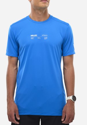 Homme portant un t-shirt de sport à manches courtes bleu vif avec un logo et un texte blanc "MILLET" sur la poitrine, un pantalon noir et une montre-bracelet blanche.