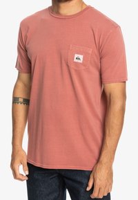 Krátký rukáv tričko v barvě nazelenalé růže, vyrobené z bavlny. Obsahuje přední kapsu s logem, kulatý výstřih a volný střih.