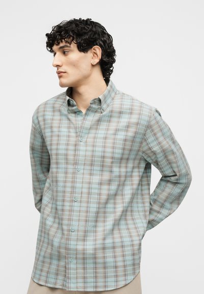 Νέος άνδρας με σγουρά σκούρα μαλλιά, φορώντας πουκάμισο καρό σε αποχρώσεις του μέντας και καφέ, στέκεται με τα χέρια πίσω από την πλάτη, κοιτάζοντας προς τα αριστερά.