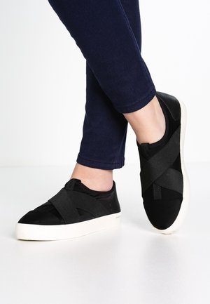 Persona con zapatillas negras sin cordones con correas elásticas cruzadas y suelas blancas, combinadas con pantalones ajustados azul oscuro.