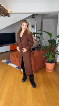 Ruskea pitkä takki, jossa vyö, jämäkät olkapäät ja napit. Päällä musta paita ja mustat housut, kuvattuna kotimaisemassa.