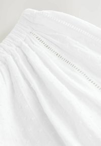 Robe blanche en tissu texturé avec des points en relief, taille élastique et bordure en dentelle décorative. Matière douce et légère avec un design drapé.
