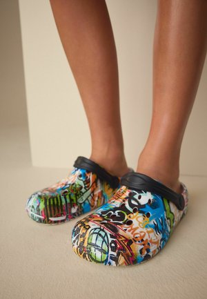 Jalad, mis kannavad värviliste graffiti-mustritega slip-on klotse, millel on mustad reguleeritavad kanna rihmad, helel põrandal ühtlase taustaga.