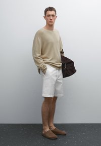 Beige striktrøje, hvide shorts og brune ruskinds slip-ons. Står med en brun taske, minimalistisk baggrund.