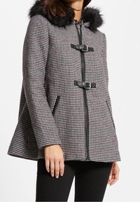 Cappotto houndstooth in bianco e nero con cappuccio foderato in pelliccia, chiusura a zip e fibbie nere. Presenta tasche laterali e un design con orlo svasato.